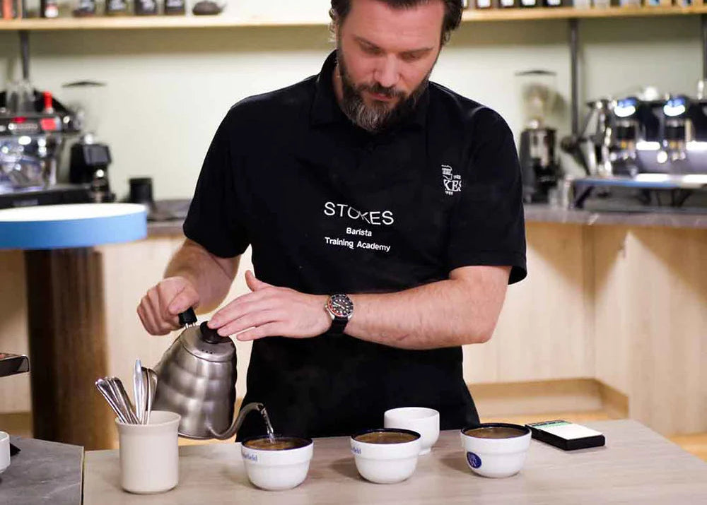 A person conducting a coffee tasting experience at a table with various coffee-related items, including a coffee grinder, scales, and cups, with the Stokes brand visible in the background.